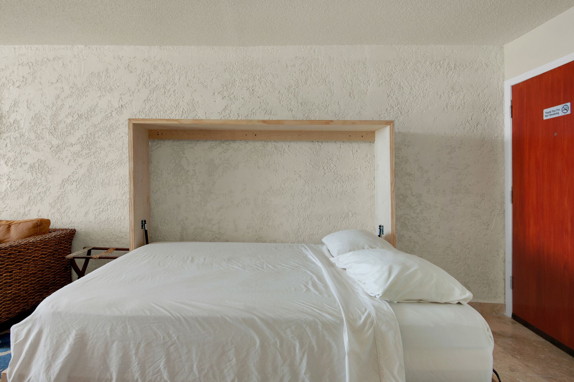 A bedroom with a wooden headboard, a bed with white bedding, a wicker chair, and a red door.