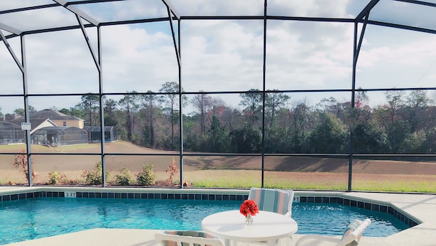 A covered outdoor swimming pool with a table and chairs, surrounded by a metal fence, and a view of a golf course and trees in the background. - 5*Luxury Villa 6BR,4BA 3Ensuits /Pool facing SE/10mins to Disney parks,free Wifi (Kissimmee)