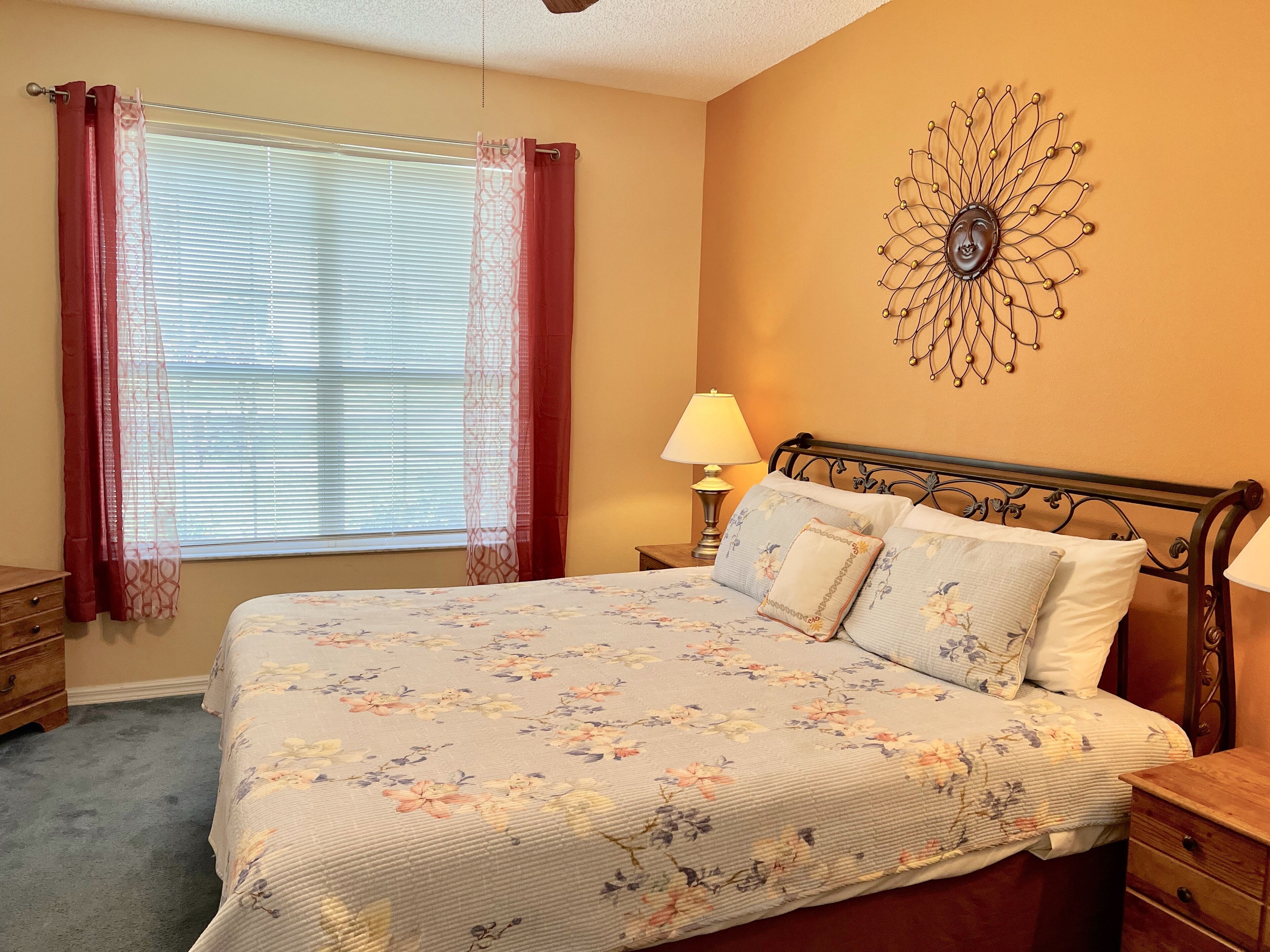 A bedroom with a bed, a nightstand, a lamp, a window with curtains, and a wall decoration.
