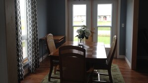 Un coin repas avec une table en bois, des chaises et un vase rempli de fleurs. Il y a deux fenêtres avec des rideaux, un lustre et une vue sur un bâtiment à l’extérieur.