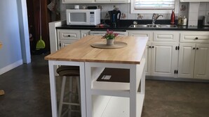 Une cuisine avec des meubles blancs, un îlot central en bois, un four à micro-ondes, une machine à café et un évier.