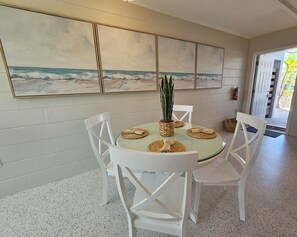 Zona pranzo con tavolo in vetro, sedie bianche e un grande dipinto a tema spiaggia sulla parete.
