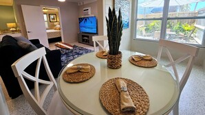 Ett modernt vardagsrum med ett glasbord, vita stolar, en tv och ett stort fönster med utsikt över omgivningen.