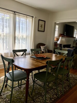 Uma área de refeições com mesa de madeira e cadeiras verdes, um tapete estampado e uma janela com cortinas leves.