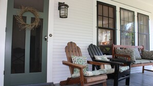 A porch with two rocking chairs and a bench, a door with a wreath, and a lantern on the wall.