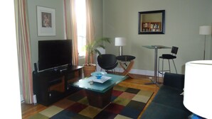 A living room with a flat-screen TV, a coffee table, a sofa, a chair, a lamp, a bookshelf, and a window with curtains. - Down Town Heritage Suite on Water Street           (Charlottetown)