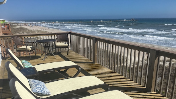 A wooden deck with lounge chairs and a table, overlooking a beach and ocean with a pier in the distance. - Panoramic Oceanfront home on Emerald Island. Close to everything!! (Emerald Isle)