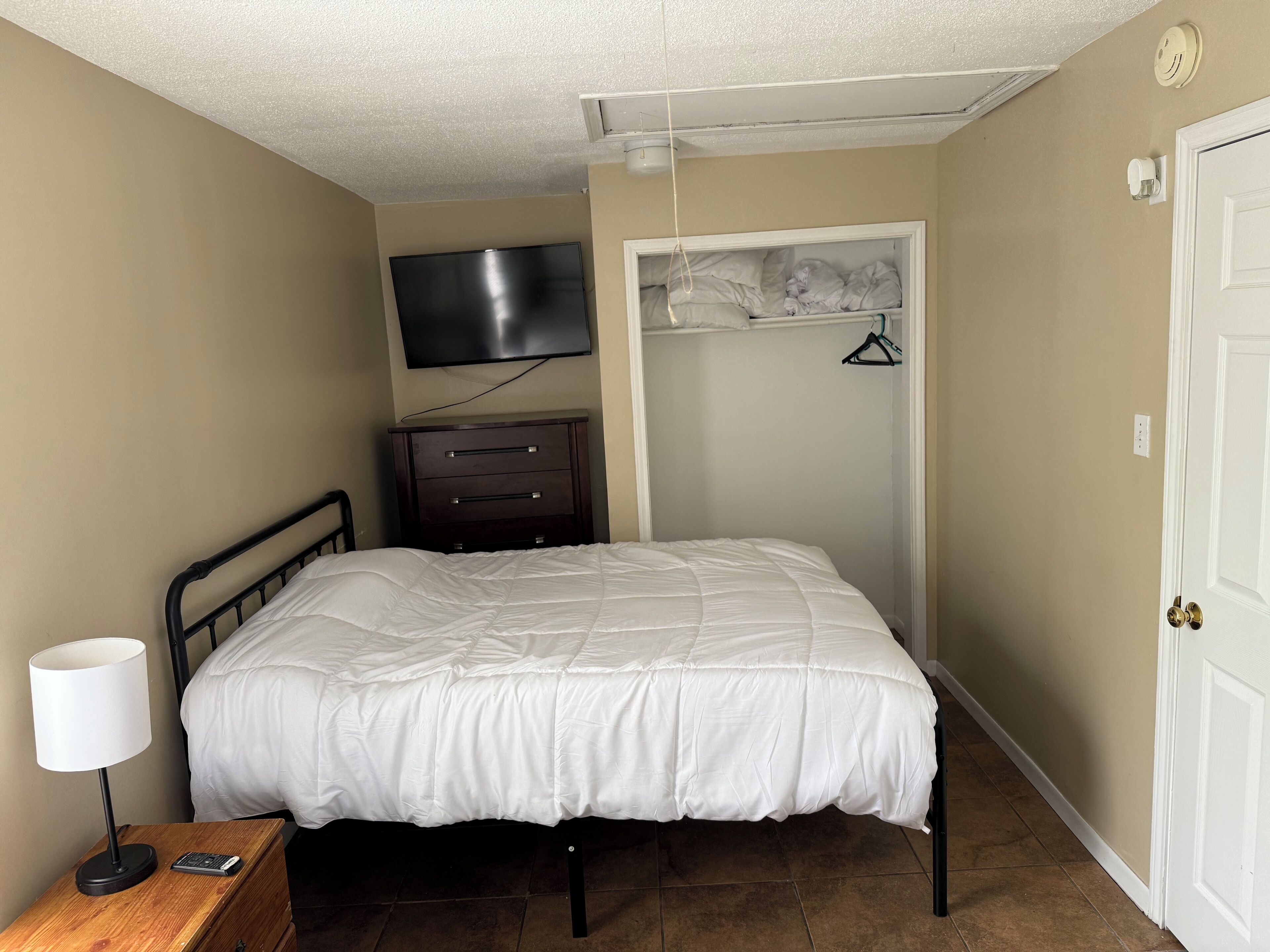 Een slaapkamer met een bed, een dressoir, een televisie aan de muur, een kledingkast met deur en een lamp op een nachtkastje.
