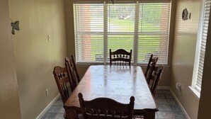 Une salle à manger avec une grande table en bois et six chaises, de grandes fenêtres équipées de stores et un ventilateur de plafond.