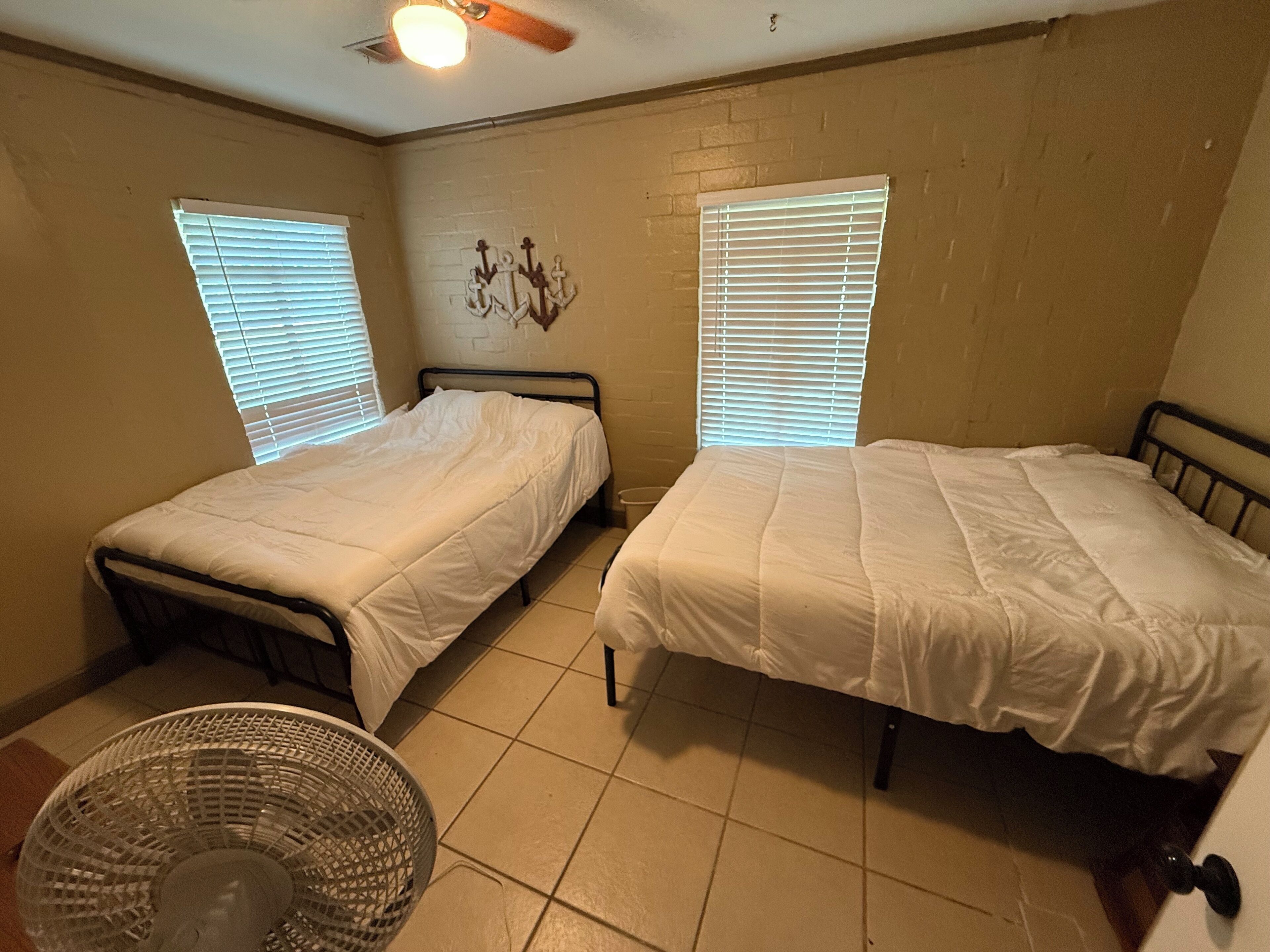 Une petite chambre avec deux lits simples, un ventilateur de plafond et deux fenêtres équipées de stores.