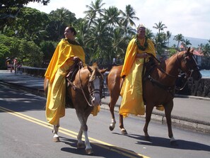 Two individuals in yellow robes riding horses on a road with palm trees and a body of water in the background. - Ocean View, Full Kitchen, Gorgeous Top Floor Suite surrounded by Tropical Garden (Kailua-Kona)