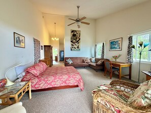 A room with a bed, a sofa, a dining table, and a chair. There is a ceiling fan, a window with curtains, and a wall with a picture.