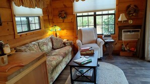 A cozy cabin interior with a floral-patterned sofa, a white armchair, a small wooden table with books, and a window with a view of greenery. - Cozy mountain cabin with amazing views, year round hot tub and pet friendly (Waynesville)