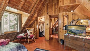 A wooden cabin with a loft bedroom, a bed with a quilt, a dresser, a chair, and a painting.
