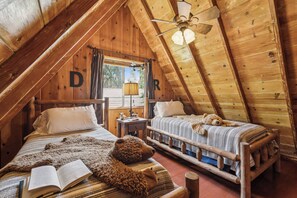 A cozy attic bedroom with wooden beams, two beds, a ceiling fan, and a window with curtains. - Rustic 1926 Lodge Style Cabin Near Lake + Spa + 3 Bonus Rooms (Lake Arrowhead)