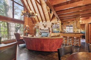A log cabin interior with a fireplace, wooden furniture, and a view of the outdoors. - Rustic 1926 Lodge Style Cabin Near Lake + Spa + 3 Bonus Rooms (Lake Arrowhead)