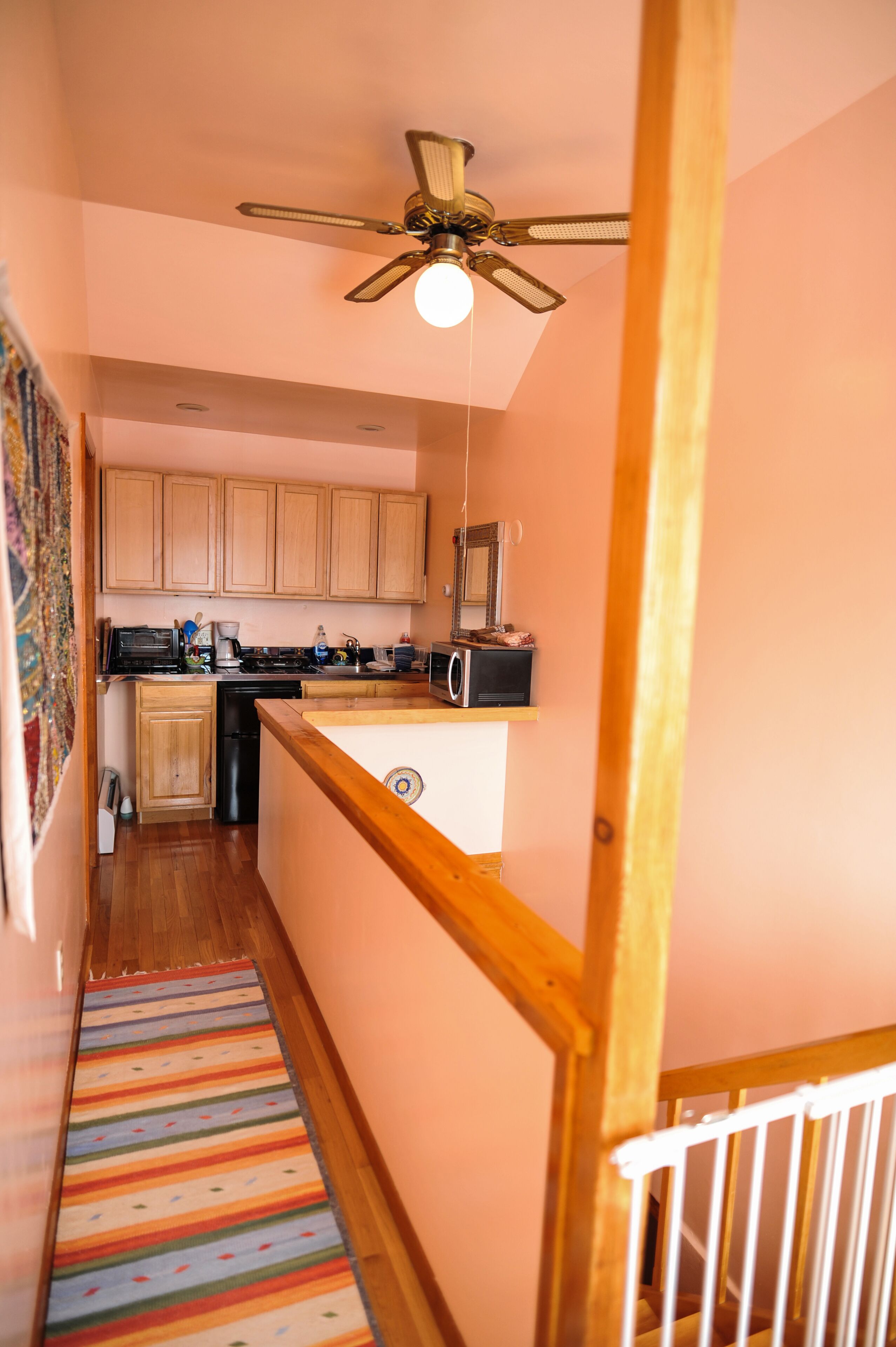 A narrow hallway with a wooden staircase, a kitchen area with wooden cabinets, a ceiling fan, and a colorful striped rug.