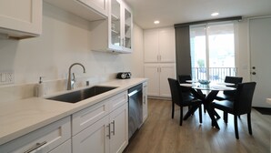 A modern kitchen with white cabinets, a stainless steel sink, and a dishwasher. There is a dining area with a round table and black chairs. - On the Beach!!  Perfect for Families.  Right on the Water (San Diego)