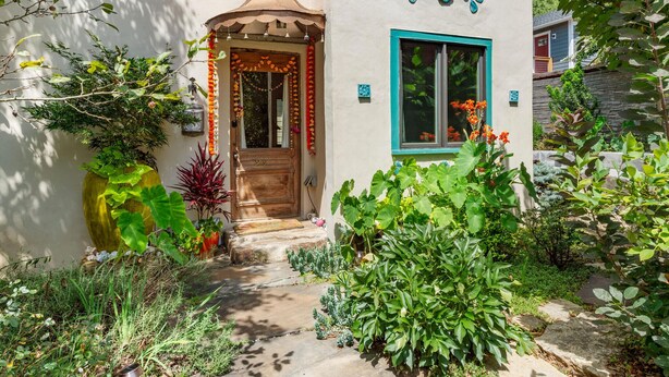 Un chemin de jardin menant à une maison dotée d’une entrée décorative, d’une grande plante en pot et d’un petit abri de jardin en arrière-plan.