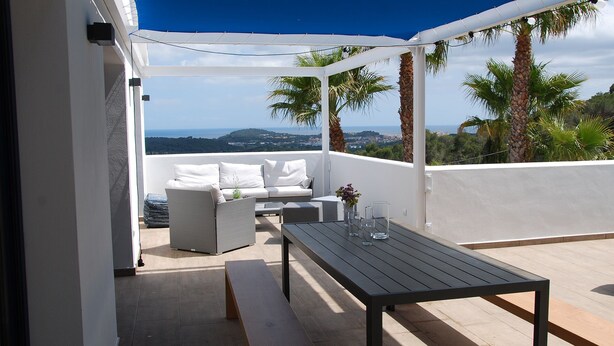 A modern outdoor seating area with a white sofa and a wooden table, overlooking a scenic landscape with palm trees and distant hills. - Modern Seaview Villa With Infinity-Pool (Palamos)