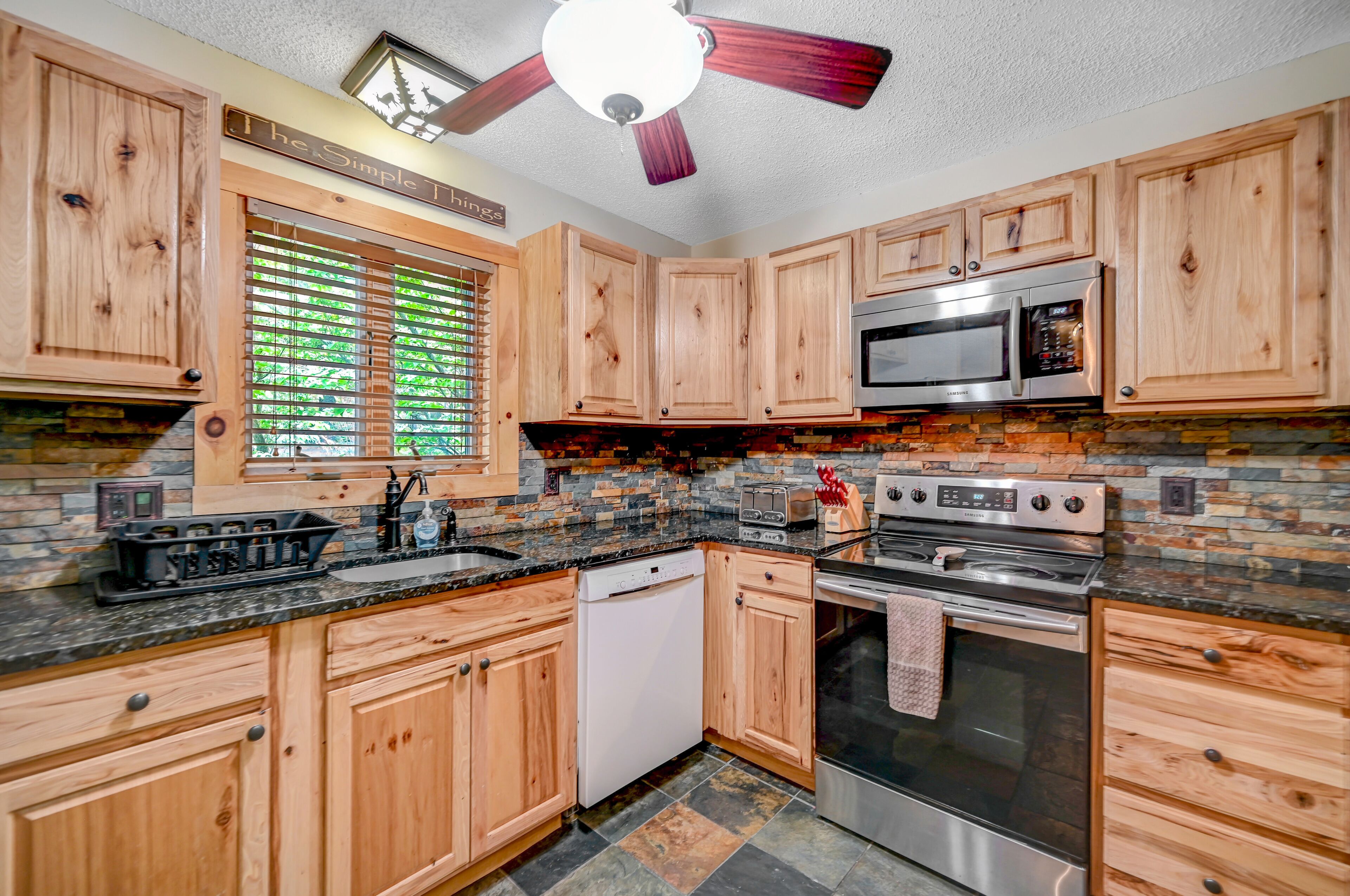A kitchen with wooden cabinets, a stone backsplash, a microwave, a stove, and a ceiling fan.