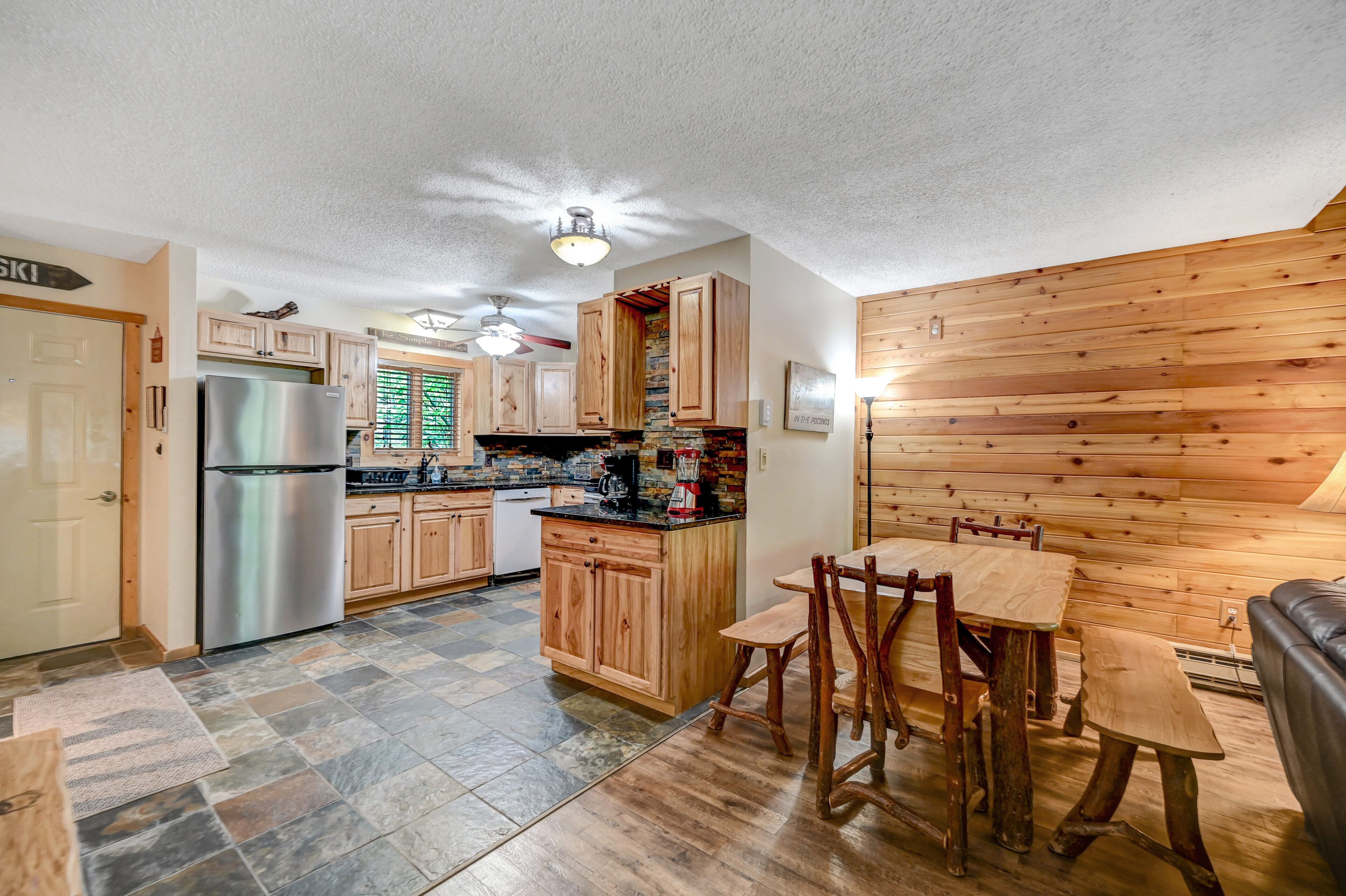 A kitchen with wooden cabinets, a refrigerator, a dining table with chairs, and a stone-tiled floor.