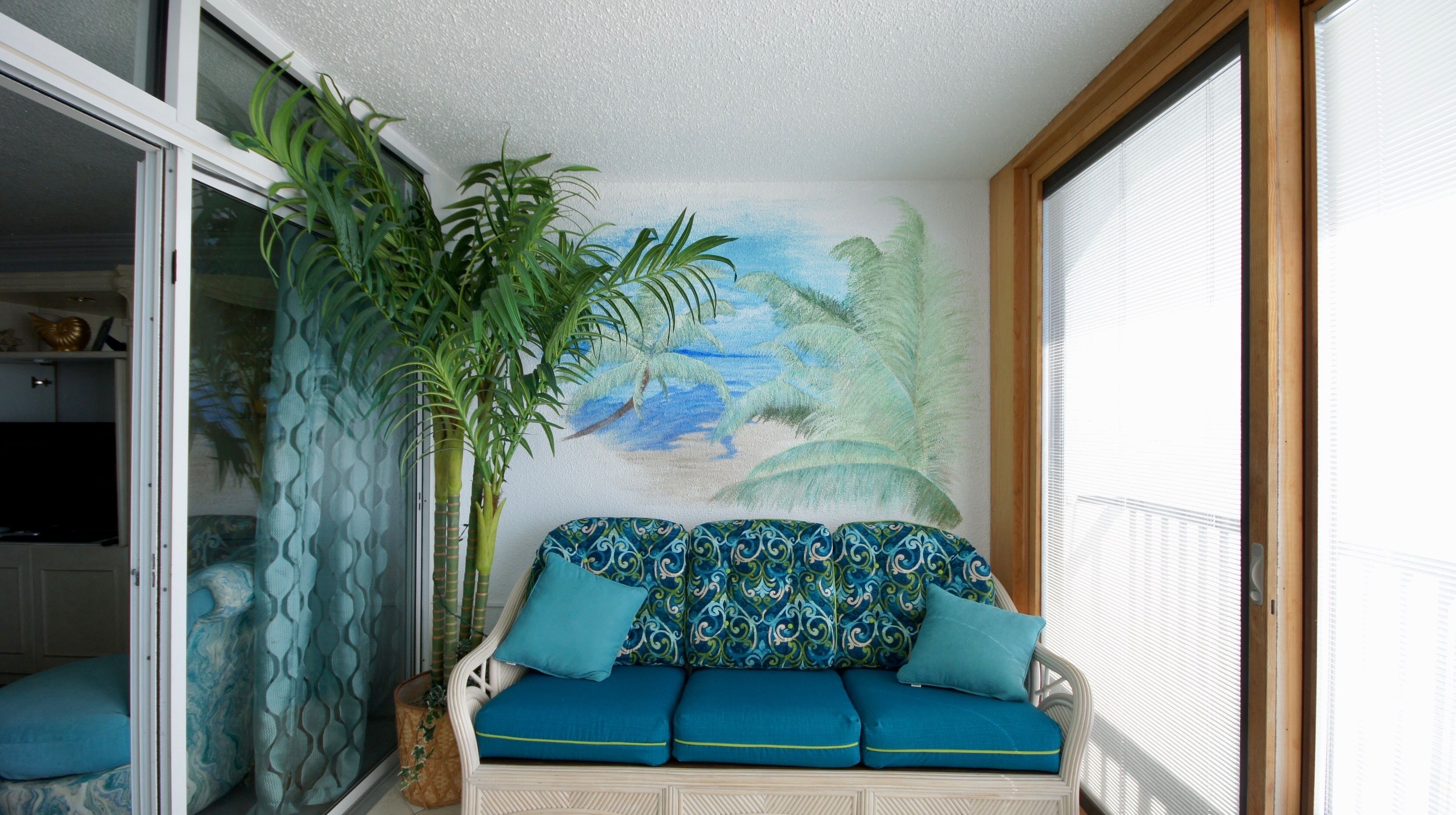 Une pièce avec un canapé en osier blanc agrémenté de coussins bleus, une grande plante en pot et une fresque murale représentant une scène de plage.