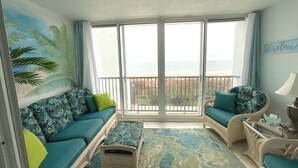 Habitación con sofá, sillón y mesita, vista al océano a través de grandes ventanales y un mural tropical en la pared.