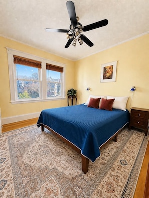 Quarto com cama, ventoinha no teto, janela, mesa de cabeceira e tapete.