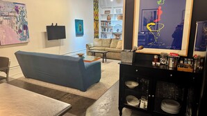 Un moderno salón con un sofá azul, una mesita de centro y un armario negro con cristalería. Hay una estantería con libros y objetos decorativos, y un cuadro abstracto grande en la pared.