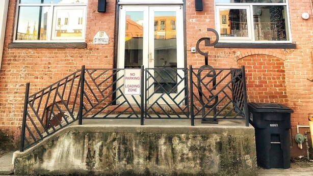 Un edificio de ladrillo con puerta de vidrio, una reja con letrero de 'No Estacionar' y un contenedor de basura negro.