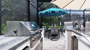 An outdoor kitchen with a stainless steel grill, a dining table with chairs, and a large umbrella providing shade.