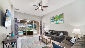A spacious living room with a sofa, coffee table, and a ceiling fan. There is a large window with a view of a pool and outdoor furniture.