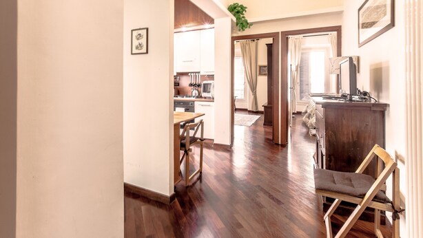 A spacious room with a wooden floor, a dining area with a table and chairs, a kitchen in the background, and a large window with curtains.
