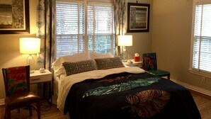 Un dormitorio con cama, dos mesitas de noche, una silla, ventilador de techo y dos ventanas con persianas.