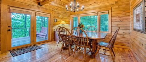 Interior de una cabaña de madera con una mesa de comedor, sillas, una araña y ventanas amplias que dan a un bosque.