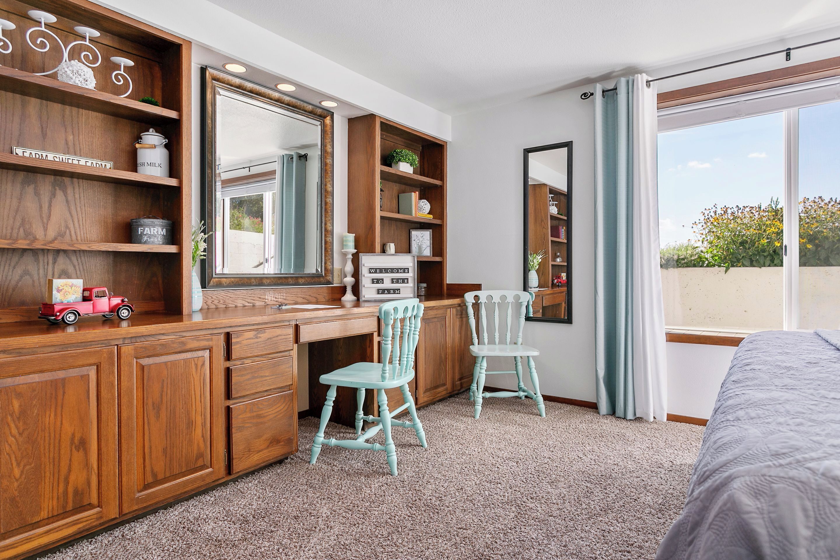 A bedroom with a wooden desk, two chairs, a large mirror, and a window with a view of greenery.
