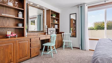 A bedroom with a wooden desk, two chairs, a large mirror, and a window with a view of greenery.