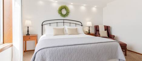 Una cama bien hecha con una manta gris, dos mesitas de noche con lámparas, un sillón de cuero y una corona verde en la pared.
