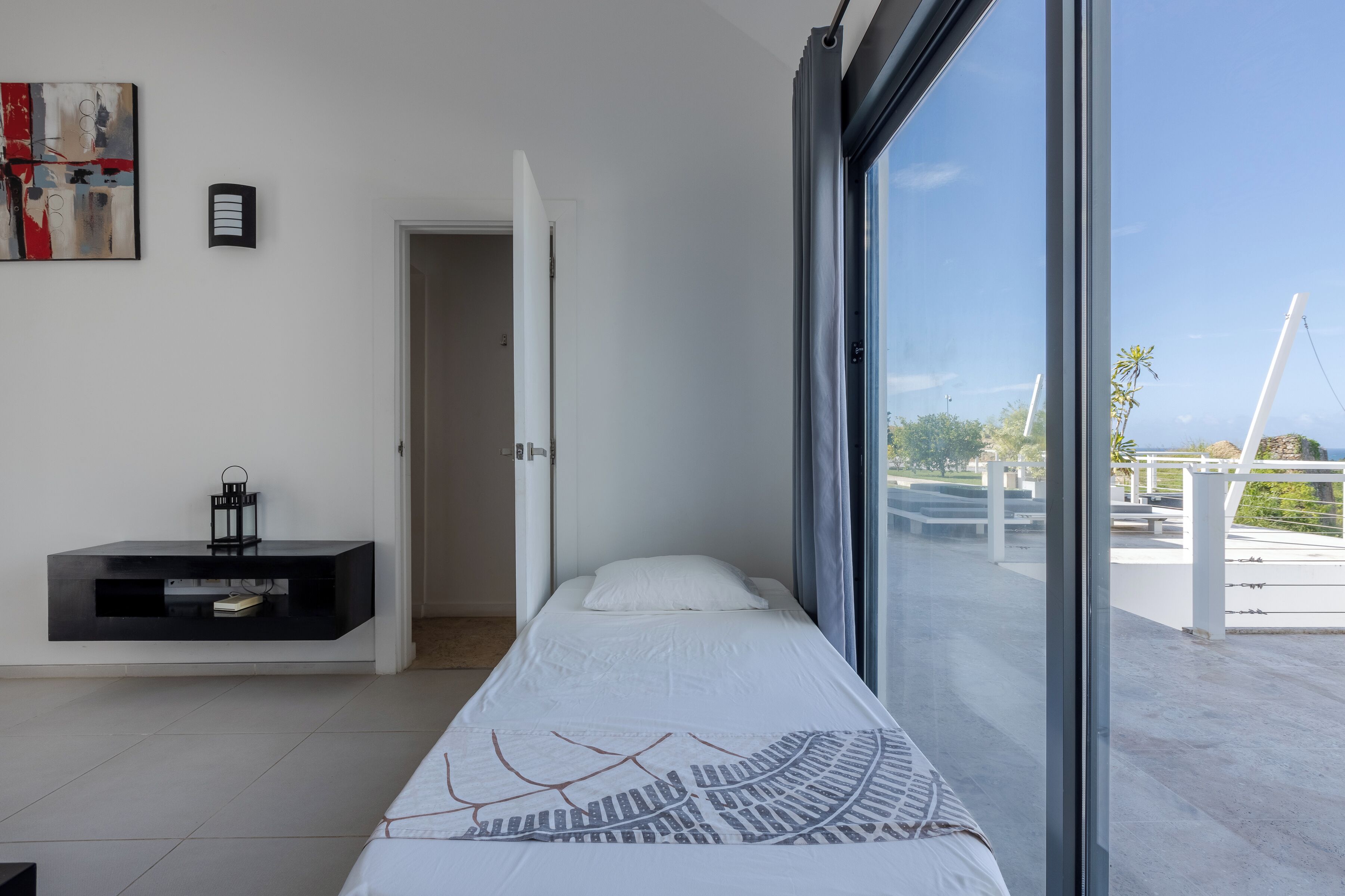 A modern bedroom with a large window, a bed with a patterned bedspread, a black console table, and a painting on the wall.