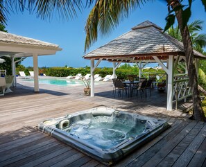 A wooden deck with a hot tub, lounge chairs, and a gazebo overlooking a pool and ocean.