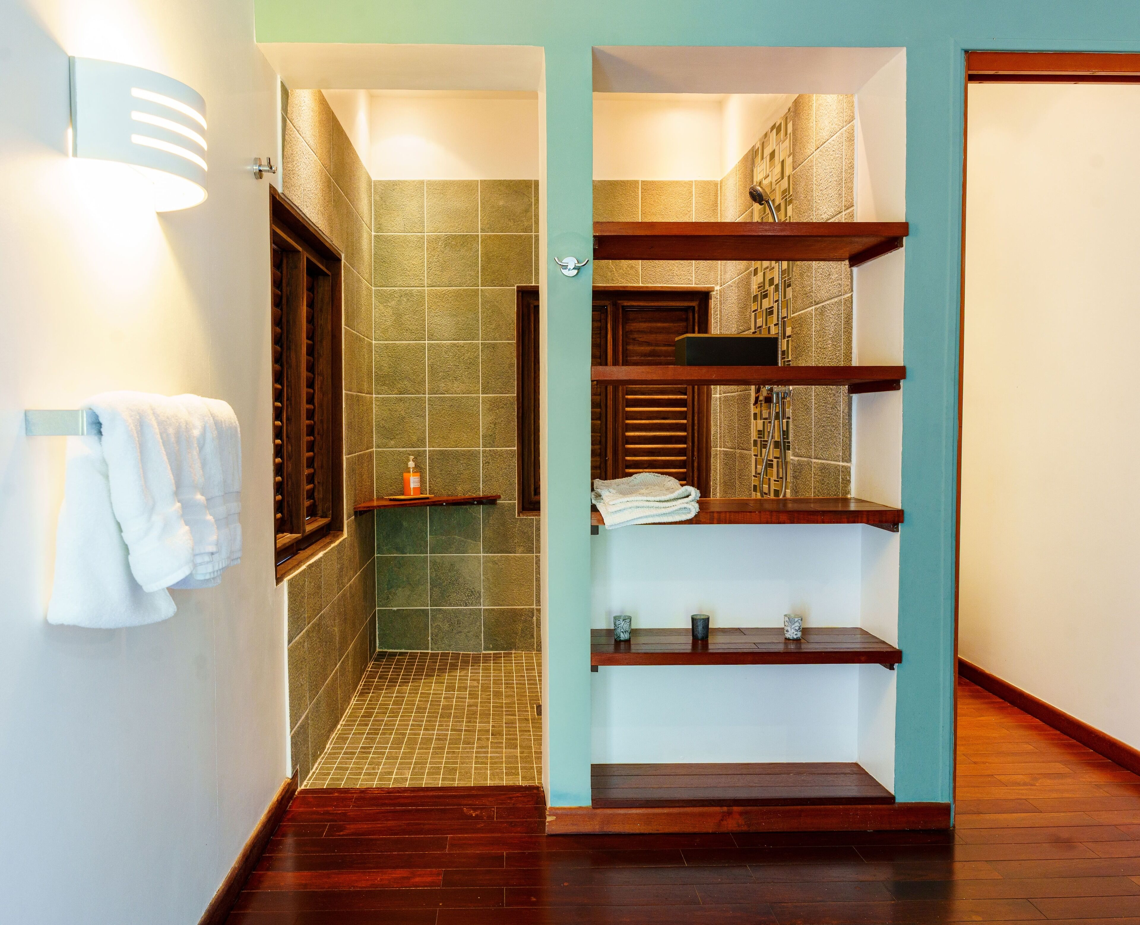 A bathroom with a shower, a wooden shelf with towels, and a tiled wall.