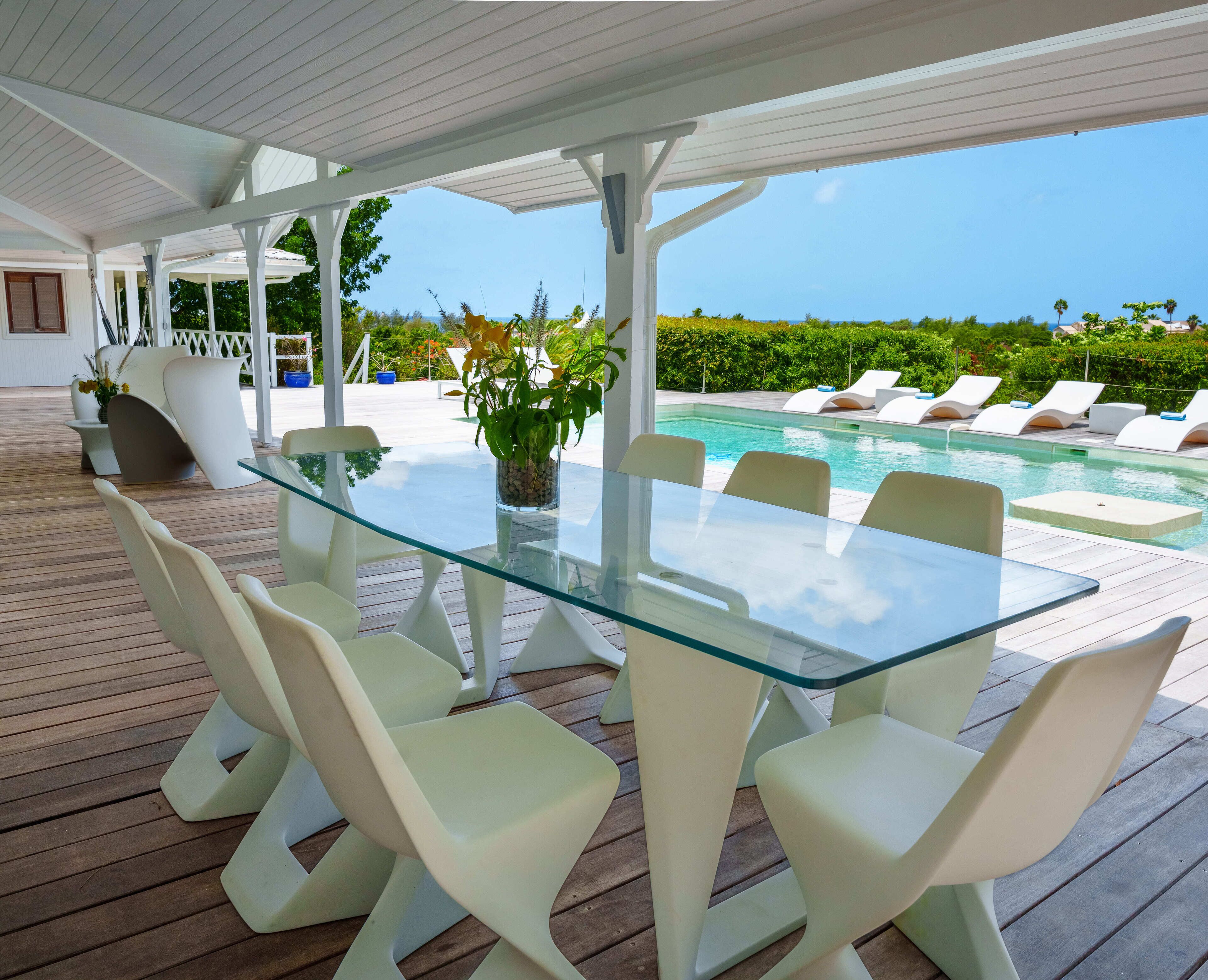 A glass-top table with white chairs, a potted plant, and a view of a pool and greenery.