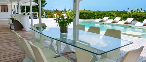 A glass-top table with white chairs, a potted plant, and a view of a pool and greenery.