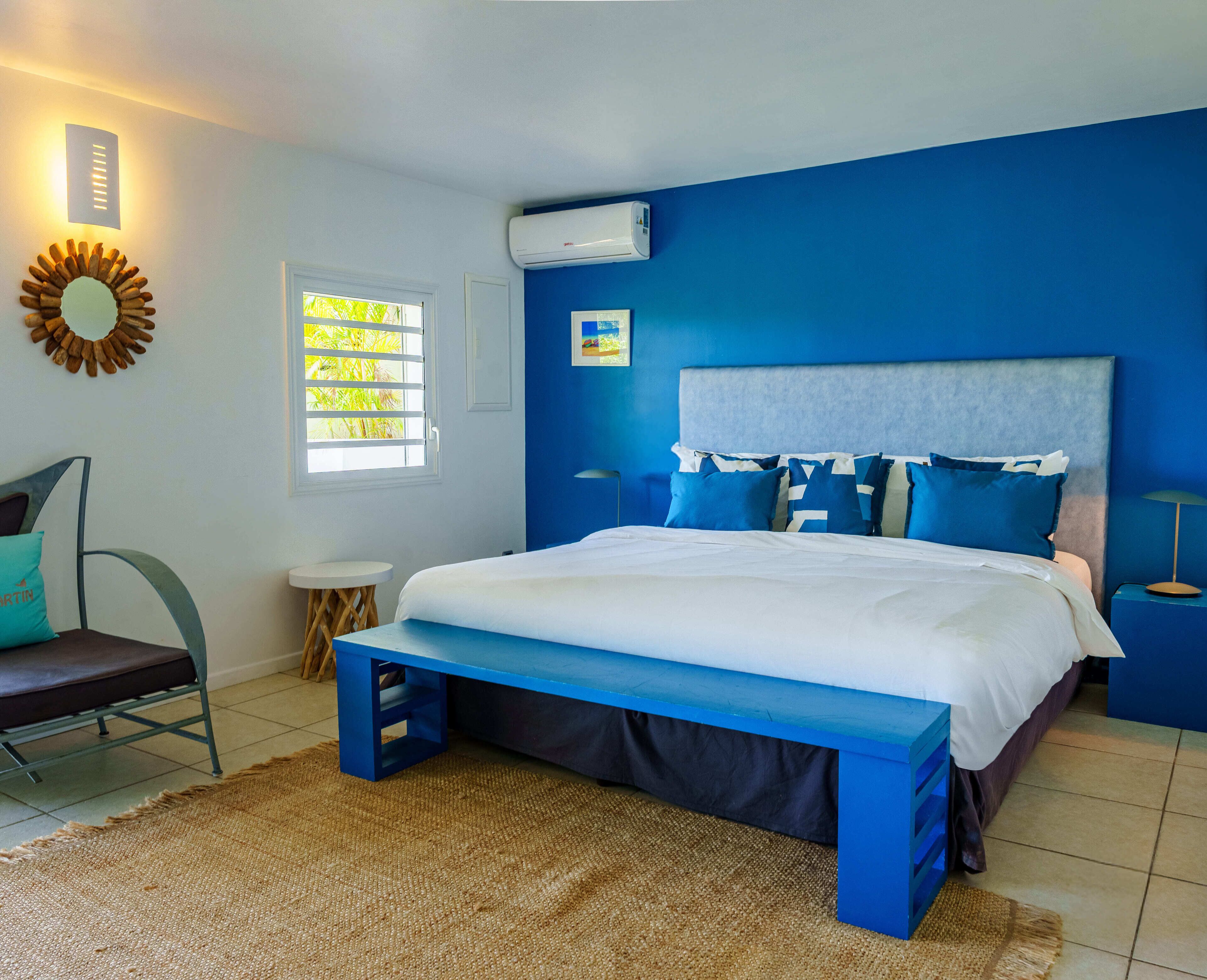 A bedroom with a bed, blue walls, a window with blinds, a wall-mounted air conditioner, and a small side table.