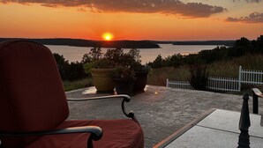Uma cadeira voltada para um pôr do sol sobre a água, com uma planta em vaso e uma cerca branca ao primeiro plano.