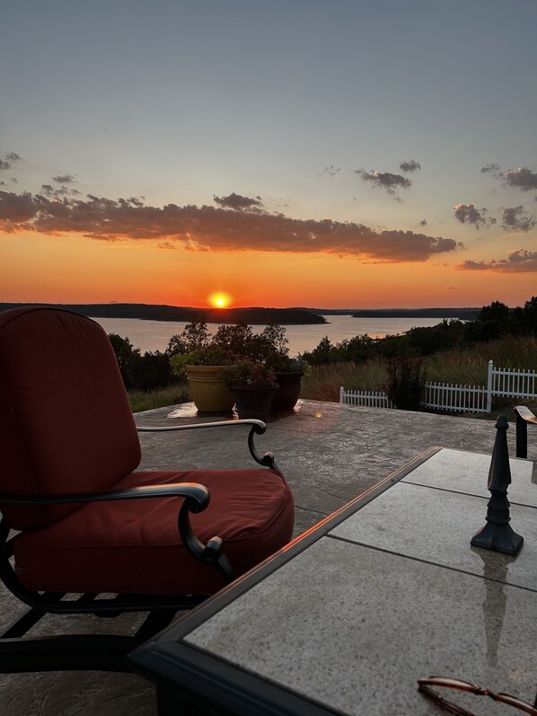 A chair facing a sunset over a body of water, with a potted plant and a white fence in the foreground.