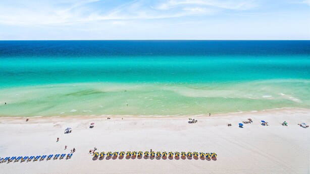 Vue aérienne d’une plage aux eaux turquoise et limpides, bordée d’un rivage sablonneux et de rangées de parasols.