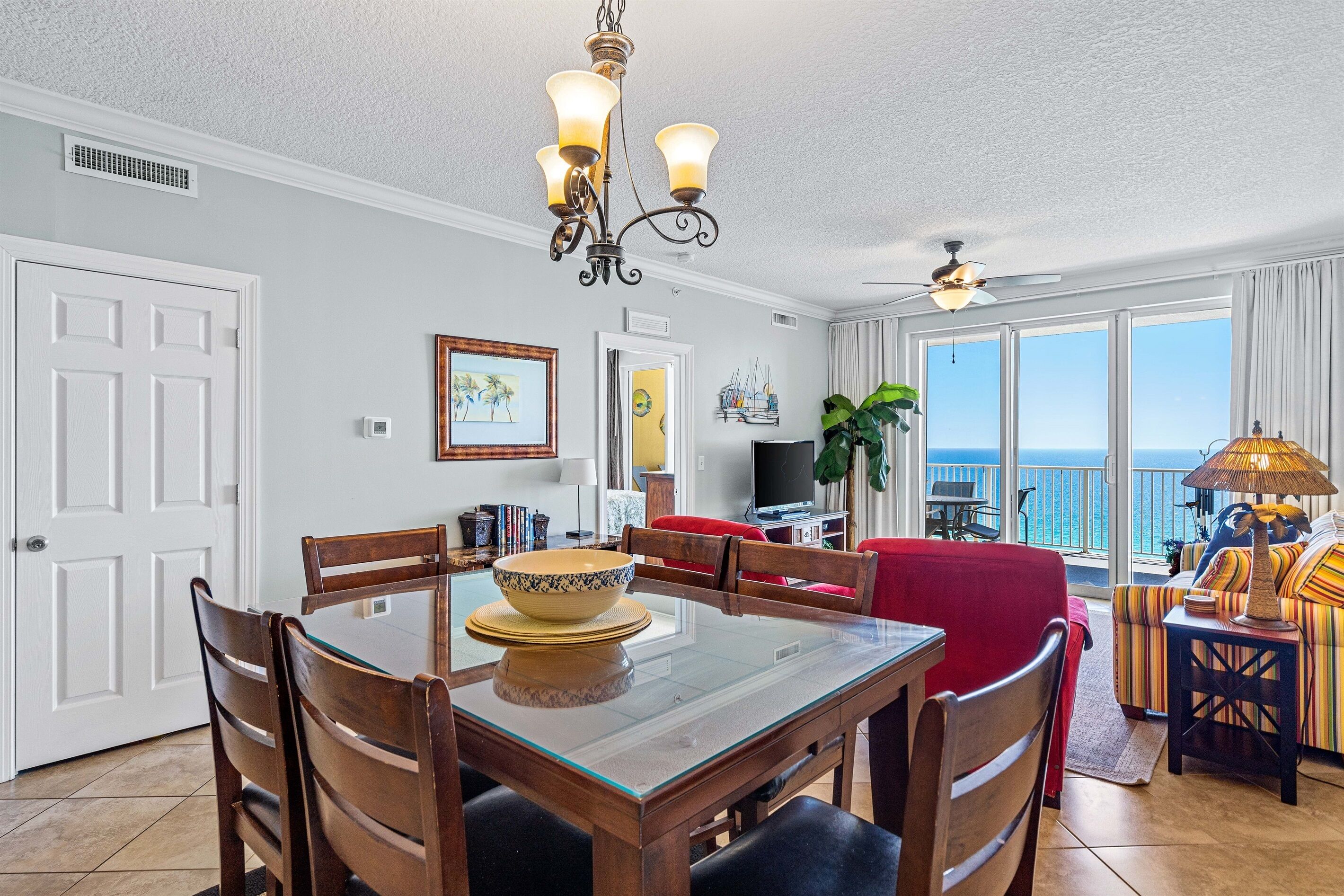 A dining area with a glass-top table, wooden chairs, and a chandelier. There is a red sofa and a TV in the background.