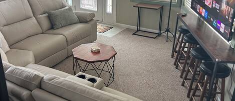A living room with a beige sectional sofa, a small round coffee table, a TV on a stand, and a bar area with stools.